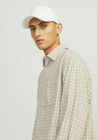 Rutete beige skjorte med en let tekstur, der har en knap-ned krave og en enkelt brystlomme. Parret med en ensfarvet hvid kasket.