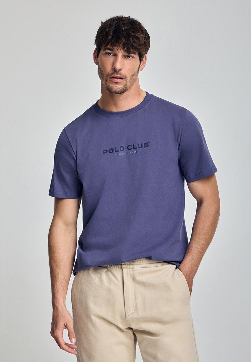 Kortärmad t-shirt med rund hals i lila bomull med en tryckt "POLO CLUB"-logotyp på bröstet. Matchas med ljusbeiga byxor.