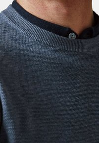 Pull bleu foncé en tricot texturé avec un col rond. Présente un col contrastant en denim marine avec un détail de bouton bleu.