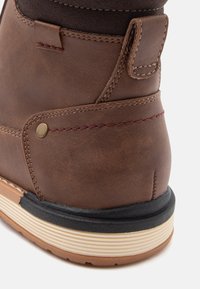 Ruskean nahkasaappaat tekstuuripinnalla, joissa on erottuva musta kumipohja ja koristeellinen punainen ommel saumoissa.