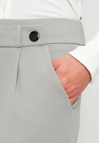 Χέρι στην τσέπη παντελονιού σε ανοιχτό γκρι χρώμα με μαύρο κουμπί, συνδυασμένο με λευκό ριμπ μακρυμάνικο τοπ.