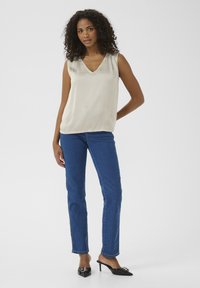 Haut sans manches crème avec un col en V, associé à un jean droit bleu et des mules à talons noirs ornées d'une boucle en argent décorative.