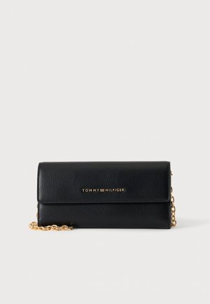 Pochette en cuir noir Tommy Hilfiger avec bandoulière chaîne dorée et lettrage logo doré sur le rabat avant sur fond blanc.