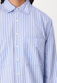 Camicia button-up azzurro chiaro con righe verticali bianche e blu scuro. Presenta due tasche sul petto, bottoni bianchi e un colletto classico.