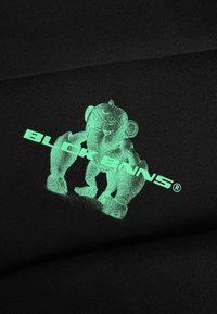 Zwart stof met een gloeien-in-het-donker afbeelding van een cartoonachtige beer en de tekst "BLOCKENNS®" in een vet, modern lettertype.