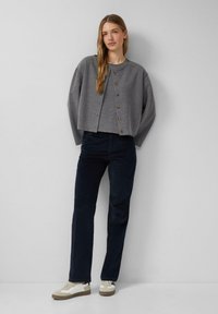 Grauer kürzerer Cardigan mit Rundhalsausschnitt und Holzknöpfen, kombiniert mit dunkelblauen Cordhosen und beigen Sneakers mit weißen Akzenten.