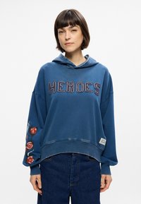 Μπλε φούτερ με κουκούλα που απεικονίζει τη λέξη "ΗΡΩΕΣ" με αντίθεση στη ραφή, κεντημένο λουλουδάτο μοτίβο στο αριστερό μανίκι και υφή με διάσταση.