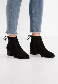 Czarne zamszowe botki sięgające kostki z wąskim noskiem i bocznym zamkiem. Niski, blokowy obcas i dekoracyjny detal z troczkiem z tyłu. Niebieskie dżinsy z poszarpanym dołem.