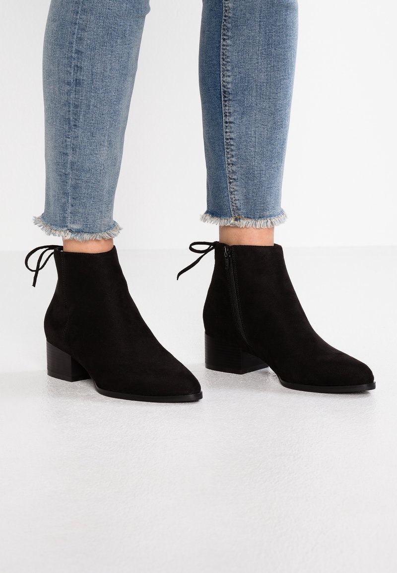 Czarne zamszowe botki sięgające kostki z wąskim noskiem i bocznym zamkiem. Niski, blokowy obcas i dekoracyjny detal z troczkiem z tyłu. Niebieskie dżinsy z poszarpanym dołem.