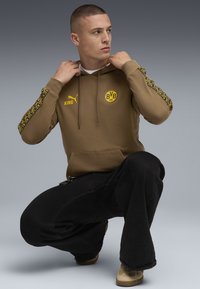 Ung mand i knæ iført en brun Puma hoodie med BVB 09 logo og sorte bukser, justerer kraven på hoodien, neutral grå studio baggrund.