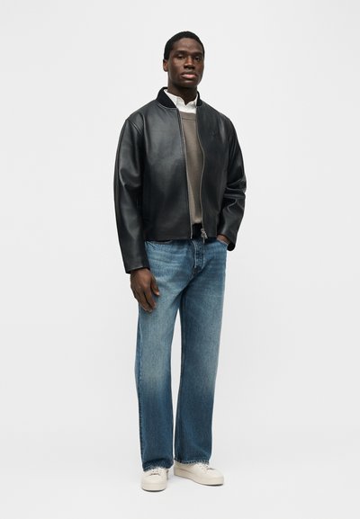 Άντρας που φοράει μαύρο δερμάτινο μπουφάν, γκρι πουλόβερ, λευκό πουκάμισο, μπλε τζιν και λευκά αθλητικά παπούτσια, στέκεται μπροστά σε λευκό φόντο.