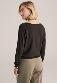 Sötétbarna pulóver laza szabással, széles nyakkivágással és hosszú ujjakkal, világos bézs nadrággal párosítva. Egyszerű dizájn, puha textúra.