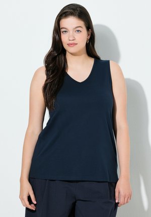 Frau mit langem dunklem Haar, die ein ärmelloses marineblaues Oberteil und passende lockere Hosen trägt, steht vor einer hellen Wand und schaut nach vorne.