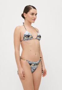 Bikini komplet s trokotnim vrhom in nastavljivimi vezavami. Primarne barve vključujejo modro, zlato in črne podrobnosti. Zlati dodatki.