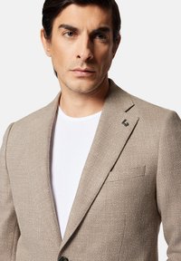 Jasnobrązowy żakiet z teksturowanym splotem, z wyłami, przednią kieszenią i małym logo na kołnierzu. Noszony na białej koszuli.