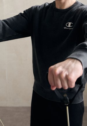 Person, der einen schwarzen Champion-Sweatshirt trägt und Griffe hält, die an Widerstandsbändern befestigt sind, während er eine Übung drinnen ausführt.