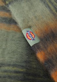 Vilnas audums zaļā krāsā ar oranžiem un brūniem rakstiem, vienā pusē ar izšūtu Dickies etiķeti. Mīksta tekstūra, abstrakts dizains.