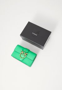 Portofel verde din piele cu accesorii din metal auriu în formă de pasăre și închidere cu buton, așezat lângă o cutie neagră etichetată "PINKO." Textură netedă, formă rectangulară.