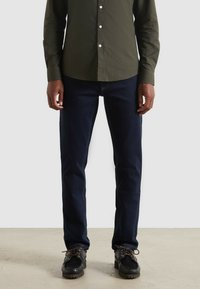 Jeans blu scuro con un design a gamba dritta, realizzati in denim, abbinati a una camicia aderente verde oliva. Vengono indossate scarpe nere con lacci.