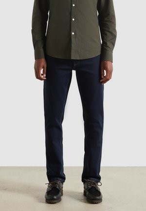 Donkerblauwe jeans met een rechte pijp, gemaakt van denim, gecombineerd met een goedzittend olijfgroen shirt. Zwarte schoenen met veters worden gedragen.