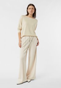 Kobieta stojąca, ubrana w beżowy luźny sweter, pasujące szerokie spodnie i spiczaste buty na tle jednolitego białego tła.