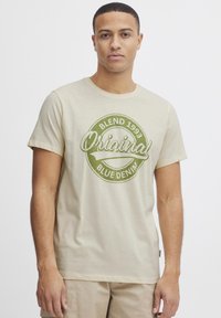 Beige bomull t-skjorte med et sirkulært grønt grafisk design med teksten "Blend 1993 Original Blue Denim." Korte ermer og rund hals.