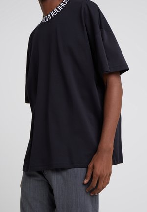 Personne portant un t-shirt noir ample avec du texte blanc sur le col et un pantalon gris, debout devant un fond uni.