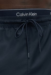 Short de bain bleu marine avec taille élastique et cordon, marque Calvin Klein visible sur la ceinture intérieure.