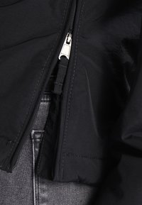 Czarna kurtka z gładkiego, teksturowanego materiału, z frontowym zamkiem błyskawicznym i srebrnym okuciem, częściowo otwarta, aby odsłonić szare dżinsy pod spodem.