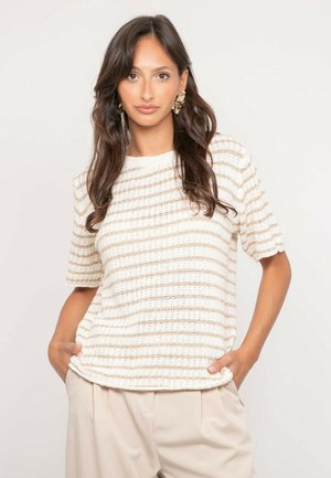 Femme aux longs cheveux bruns portant des boucles d'oreilles en feuille d'or, un haut en maille rayé crème et beige, et un pantalon beige clair les mains dans les poches.