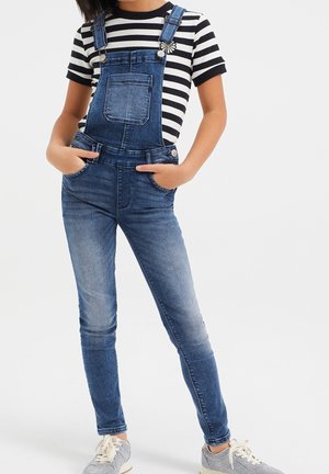 Jeans-Latzhose mit verstellbaren Trägern, Vordertasche und blauem Waschung, kombiniert mit einem schwarz-weiß gestreiften kurzärmligen Shirt und grauen Schuhen.