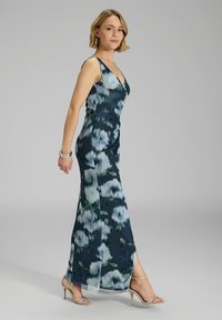 Floral Maxi-Kleid in Dunkelblau mit blauen und grünen Mustern, mit V-Ausschnitt, ärmellosem Design und seitlichem Schlitz. Getragen mit metallischen High Heels.