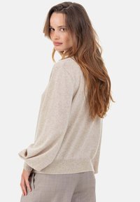 Beiger Strickpullover mit langen, lockeren Ärmeln und einem gerippten Saum. Weiche Textur mit feinem, gesprenkeltem Muster überall.