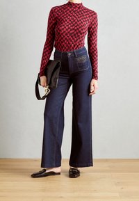 Jeans a vita alta in denim blu a gamba larga con dettagli di tasche borchiate, abbinati a un top rosso a maniche lunghe con motivo e scarpe nere senza lacci.