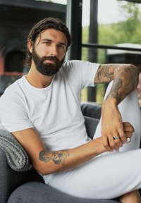 Hvid t-shirt, tatovering på armen, siddende stilling på en grå stofsofa, mørk skæg, langt hår, holder hænderne med en sølvring.