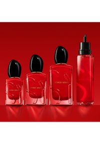 Quattro flaconi di profumo Giorgio Armani rossi di dimensioni diverse con tappi neri, disposti in fila su uno sfondo rosso.