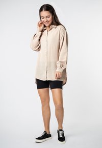 Lichtbeige langslavende shirt van een textuur met een gekreukeld stof, met knoopsluiting en een losse fit, gecombineerd met zwarte fietsbroek en zwarte sneakers.