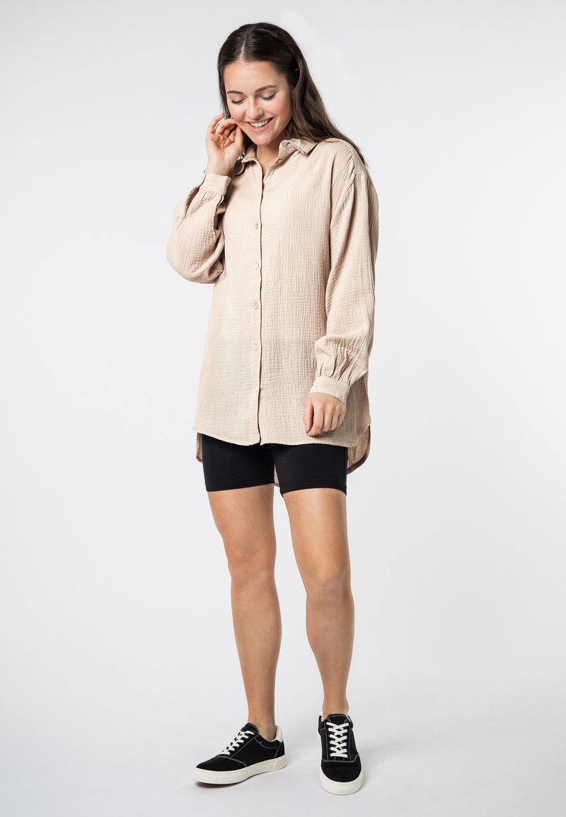 Lichtbeige langslavende shirt van een textuur met een gekreukeld stof, met knoopsluiting en een losse fit, gecombineerd met zwarte fietsbroek en zwarte sneakers.
