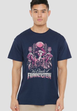 Young man wearing navy blue T-shirt with pink graphic of the Bride of Frankenstein and text below the figure.