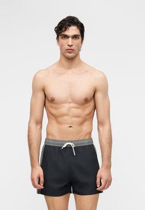 Jeune homme aux cheveux courts et foncés, à la carrure musclée, portant un short de bain noir avec une taille grise et un cordon blanc, debout devant un fond gris.