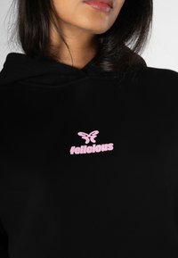 Musta huppari pehmeällä tekstuurilla, jossa on keskellä vaaleanpunainen perhoslogo ja sana "felicious" rohkealla vaaleanpunaisella kirjasimella.