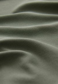 Olivgrüner Stoff, der sanfte, strukturierte Wellen mit einer glatten Oberfläche zeigt. Das Material wirkt leicht und flexibel und weist subtile Muster auf.