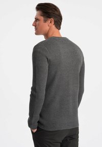 Grått texturerat stickat tröja med rund halsringning. Bakifrån syns långa ärmar och en avslappnad passform, tillsammans med svarta byxor.