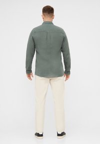 Camicia di lino verde, maniche lunghe, polsini con bottoni, dettaglio sullo yoke posteriore, abbinata a pantaloni crema e sneakers nere su uno sfondo semplice.