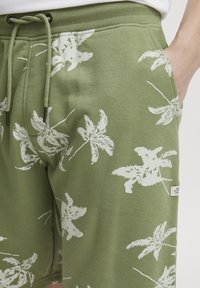 Pantaloni corti verdi con una texture morbida, caratterizzati da motivi di foglie tropicali bianche e cordoncino regolabile. Inclusi tasche laterali e un piccolo etichetta.