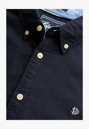 Dunkel marineblaues Hemd mit cremefarbenen Knöpfen, geknöpftem Kragen, weiß besticktem Logo auf der Brust und blauem Innenetikett am Kragen.