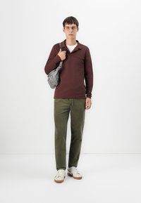 Tânăr purtând ochelari, cămașă lungă de culoare vinie, pantaloni de catifea verzi măslinii și adidași albi, ținând o curea de poșetă gri.