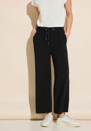 Personne portant un pantalon large noir avec taille à cordon, un T-shirt blanc et des baskets blanches, debout contre un mur beige.