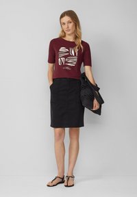 Chemise bordeaux à manches courtes avec un design graphique, associée à une jupe noire à mi-mollet et des sandales noires. Comprend un sac tissé noir.