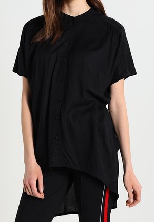 Kobieta w luźnej czarnej koszuli na krótki rękaw z guzikami, o asymetrycznym, wydłużonym tylnym kroju oraz czarnych spodniach z czerwono-białymi paskami po bokach.